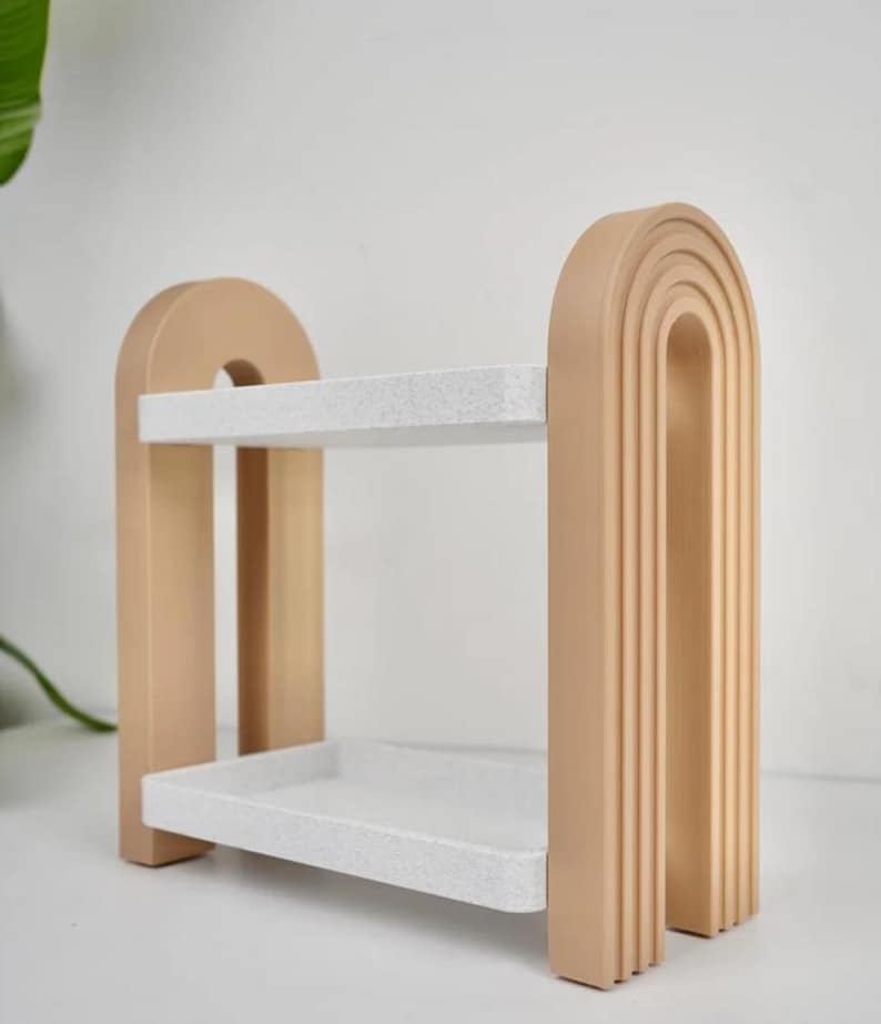 May include: A two-tiered decorative shelf in a neutral color palette. The shelf features a light beige frame with a rounded arch design and two white speckled shelves. The design is modern and minimalist, suitable for displaying small items.