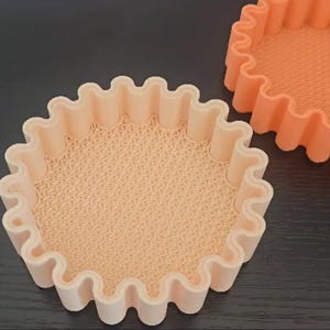 May include: Two peach-colored, round baking molds with a wavy edge. The interior of the molds has a textured, dotted pattern. One mold is in the foreground, and another is partially visible in the background.