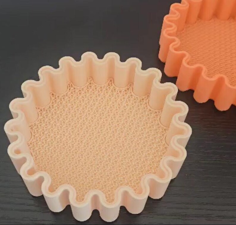 May include: Two peach-colored, round baking molds with a wavy edge. The interior of the molds has a textured, dotted pattern. One mold is in the foreground, and another is partially visible in the background.