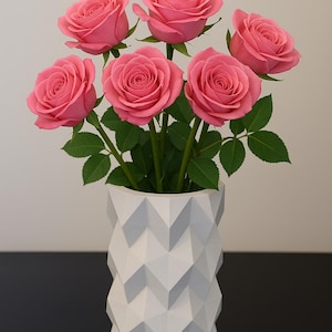 Puede incluir: Un ramo de rosas rosas en un jarr&oacute;n geom&eacute;trico blanco. Las rosas tienen m&uacute;ltiples capas de p&eacute;talos y tallos verdes. El jarr&oacute;n tiene un dise&ntilde;o moderno y angular. El arreglo est&aacute; sobre un fondo neutro.