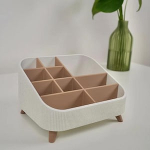 Pode incluir: Um organizador branco e castanho com vários compartimentos. O organizador tem um exterior texturizado e é elevado por pequenas pernas castanhas. Os compartimentos internos são castanhos. Um vaso de vidro verde com uma planta está no fundo.