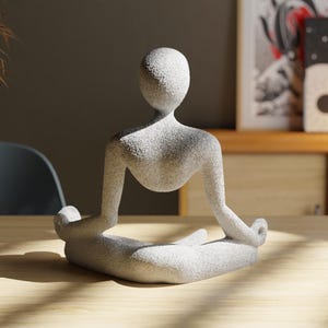 May include: A light gray stone sculpture of a person in a seated yoga pose. The figure has a smooth, rounded head and body with arms and legs in a meditative position. The sculpture is on a wooden surface.
