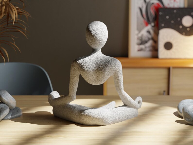 May include: A light gray stone sculpture of a person in a seated yoga pose. The figure has a smooth, rounded head and body with arms and legs in a meditative position. The sculpture is on a wooden surface.