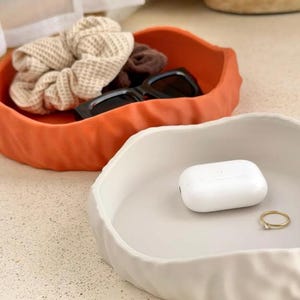 May include: Two irregular, round, shallow trays with a textured, rippled exterior. One tray is matte orange, containing a beige waffle-knit scrunchie, a brown scrunchie, and black sunglasses. The other tray is matte white, holding a white earbud case and a gold ring.