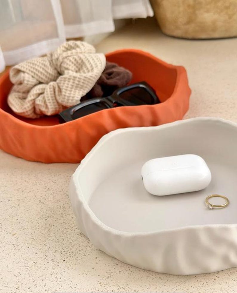 May include: Two irregular, round, shallow trays with a textured, rippled exterior. One tray is matte orange, containing a beige waffle-knit scrunchie, a brown scrunchie, and black sunglasses. The other tray is matte white, holding a white earbud case and a gold ring.