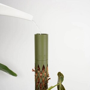 May include: An olive green plant support pole with a diamond-patterned lower section filled with brown moss. Water is being poured into the top of the pole from a white watering can. The text "moss buddy" is visible on the pole.