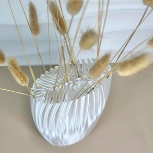 May include: A white, oval-shaped vase with a textured, ridged design, holding dried, tan-colored bunny tail grass. The vase is mounted on a wall, creating a minimalist and modern aesthetic. The arrangement is suitable for home decor.
