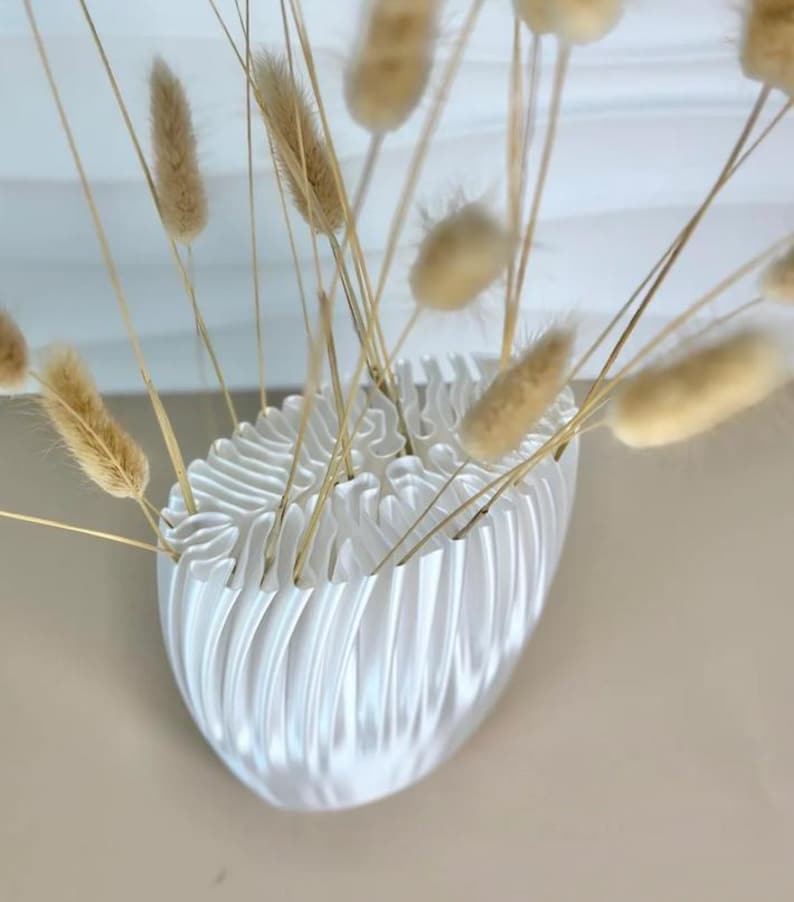 May include: A white, oval-shaped vase with a textured, ridged design, holding dried, tan-colored bunny tail grass. The vase is mounted on a wall, creating a minimalist and modern aesthetic. The arrangement is suitable for home decor.