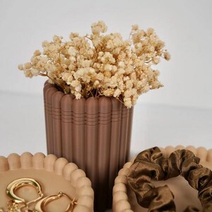 May include: A brown fluted cylindrical vase filled with dried cream-colored baby's breath flowers, positioned between two matching beige fluted containers. One container holds gold hoop earrings, and the other holds a dark brown satin scrunchie.