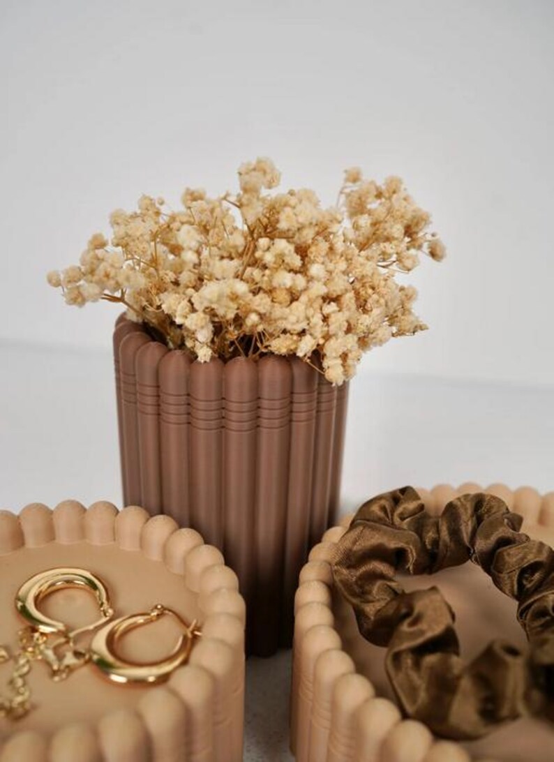 May include: A brown fluted cylindrical vase filled with dried cream-colored baby's breath flowers, positioned between two matching beige fluted containers. One container holds gold hoop earrings, and the other holds a dark brown satin scrunchie.