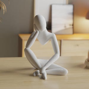May include: A white abstract sculpture of a seated figure with a textured surface. The figure is depicted in a contemplative pose, resting its head on one hand with the arm supported by a bent knee. The design features smooth, flowing lines.