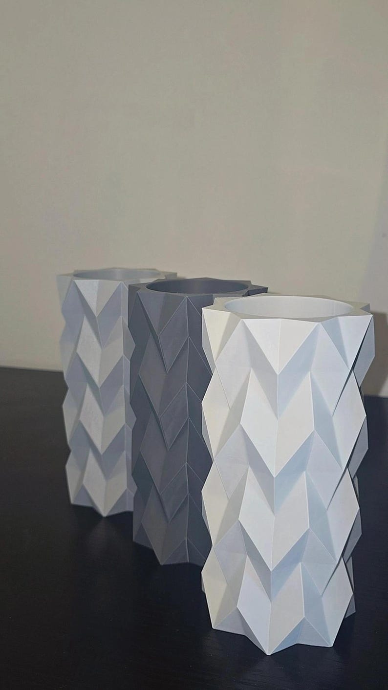 Puede incluir: Tres jarrones geom&eacute;tricos en tonos grises y blancos. Cada jarr&oacute;n tiene un patr&oacute;n de chevr&oacute;n &uacute;nico, creando una superficie texturizada. Los jarrones son cil&iacute;ndricos con una parte superior cuadrada, adecuados para la decoraci&oacute;n del hogar.