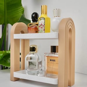 May include: A two-tiered, beige and white shelf unit displaying various perfume bottles. The shelf has an arched design and is made of wood and a speckled white material. The perfumes are in various shapes and colors, including yellow, clear, and orange.