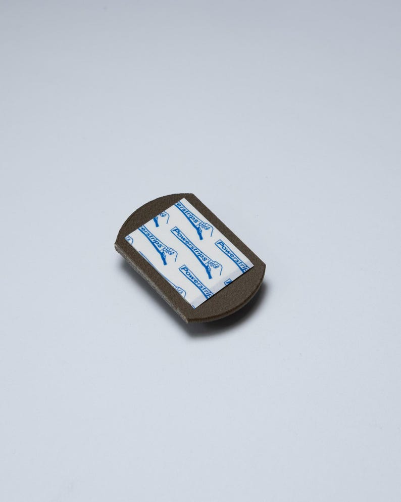 May include: A small, dark brown, semi-circular adhesive mounting piece with a flat top edge. The centre features a white rectangular adhesive pad with blue text that reads Powerstrips and includes a small icon of a hand pulling a tab.