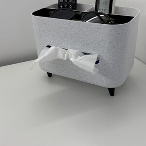 Op de afbeelding: Een lichtgrijze tissuebox met een rechthoekige vorm en zwarte poten. De box bevat een afstandsbediening, een kleine digitale thermometer en een zonnebril. Witte tissues worden uit een gleuf aan de voorkant getrokken.