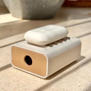 Op de afbeelding: Een rechthoekig wit zeepbakje met een gestructureerd bovenoppervlak en een glad beige eindpaneel met een ronde uitsparing. Een vast wit stuk zeep ligt bovenop het bakje. Het zeepbakje heeft een strak, functioneel ontwerp met afgeronde hoeken.