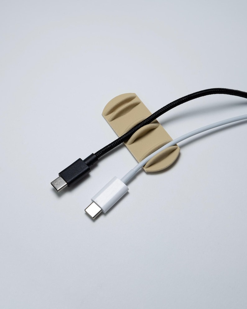 May include: A beige silicone cable organiser featuring three parallel slots designed to hold cords in place. One black braided cable and one white cable are currently secured within the slots, with their respective USB-C connectors visible.