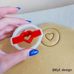 May include: A white circular cookie cutter with a red heart-shaped center insert, positioned above a piece of rolled-out dough that has a circular impression with a heart cutout. The text @Eyf_design is visible in the bottom right corner.