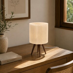 Puede incluir: Una l&aacute;mpara de mesa con una pantalla cil&iacute;ndrica plisada de color blanco roto, sostenida por tres patas de madera de color marr&oacute;n oscuro. La l&aacute;mpara emite una luz suave y c&aacute;lida e incluye un cable de alimentaci&oacute;n blanco en la base del escritorio.