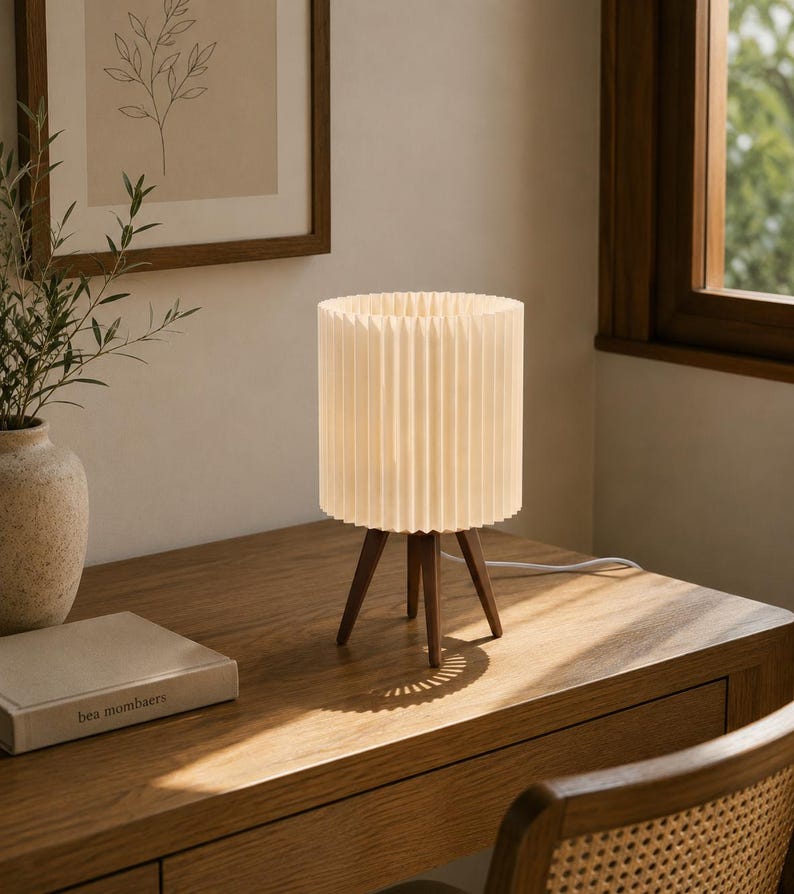 Puede incluir: Una l&aacute;mpara de mesa con una pantalla cil&iacute;ndrica plisada de color blanco roto, sostenida por tres patas de madera de color marr&oacute;n oscuro. La l&aacute;mpara emite una luz suave y c&aacute;lida e incluye un cable de alimentaci&oacute;n blanco en la base del escritorio.