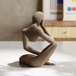 May include: A brown, textured sculpture of a person in a seated, thoughtful pose. The figure is resting their chin on one hand, with the other arm on their knee. The sculpture has a rough, granular surface, possibly made of stone or a similar material.