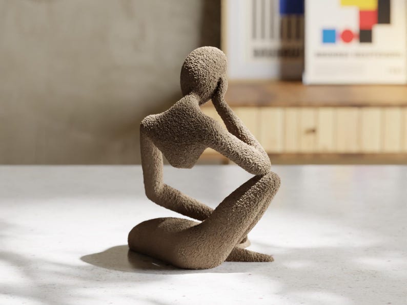 May include: A brown, textured sculpture of a person in a seated, thoughtful pose. The figure is resting their chin on one hand, with the other arm on their knee. The sculpture has a rough, granular surface, possibly made of stone or a similar material.
