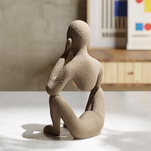 May include: A beige, textured sculpture of a seated human figure in a contemplative pose. The figure has a rounded head, a curved back, and bent knees, with one hand resting near the face. The surface has a granular, sand-like finish throughout.