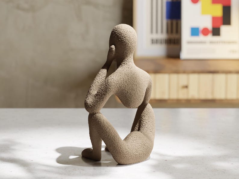May include: A beige, textured sculpture of a seated human figure in a contemplative pose. The figure has a rounded head, a curved back, and bent knees, with one hand resting near the face. The surface has a granular, sand-like finish throughout.