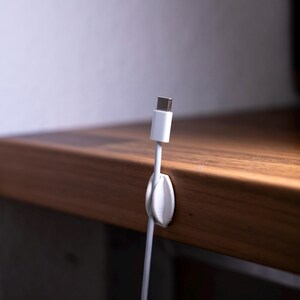 May include: A white plastic cable organiser clip attached to the edge of a wooden surface, holding a white USB-C charging cable in an upright position. The clip features a rounded, leaf-like design that secures the cable cord firmly in place.
