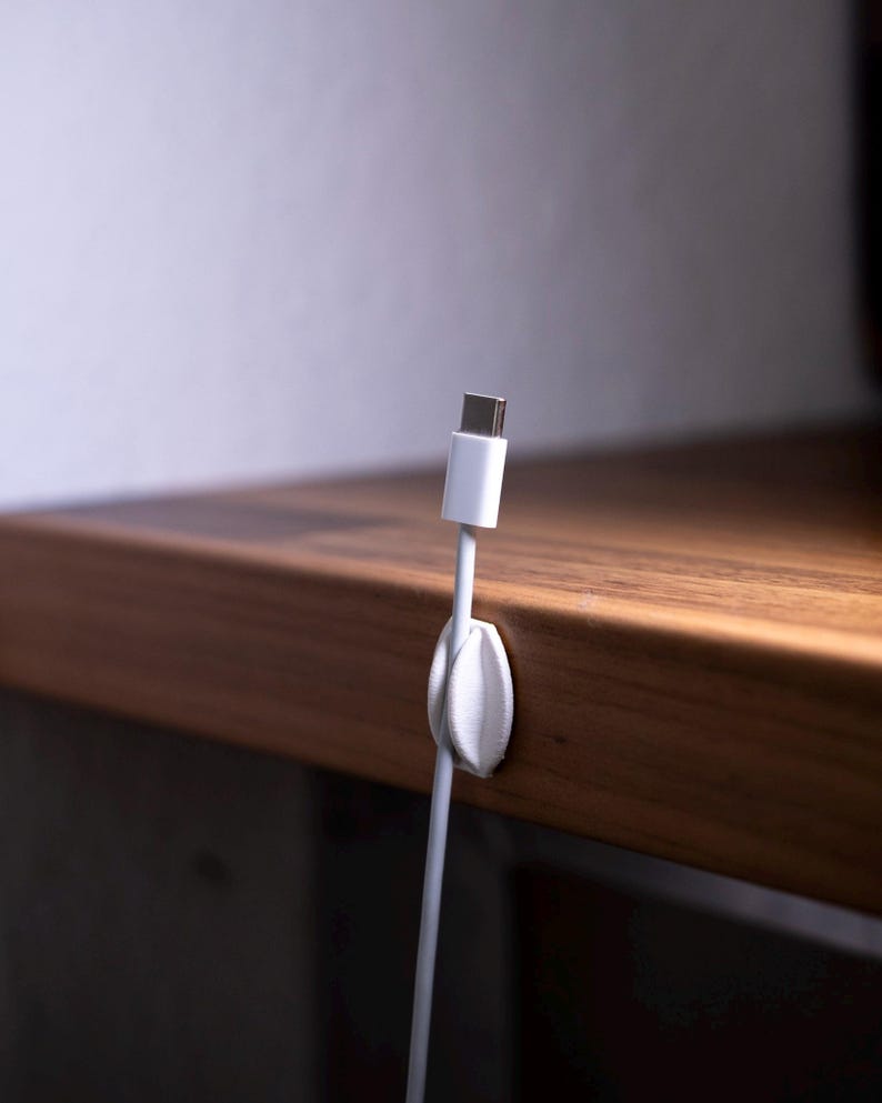 May include: A white plastic cable organiser clip attached to the edge of a wooden surface, holding a white USB-C charging cable in an upright position. The clip features a rounded, leaf-like design that secures the cable cord firmly in place.