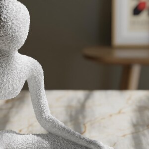 May include: A white, textured sculpture of a seated figure with an arm extended. The sculpture has a rough, granular surface and is set against a neutral background. The figure's form is simplified and abstract.