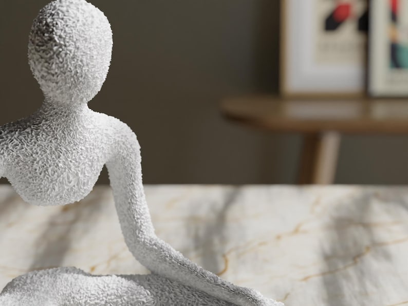 May include: A white, textured sculpture of a seated figure with an arm extended. The sculpture has a rough, granular surface and is set against a neutral background. The figure's form is simplified and abstract.