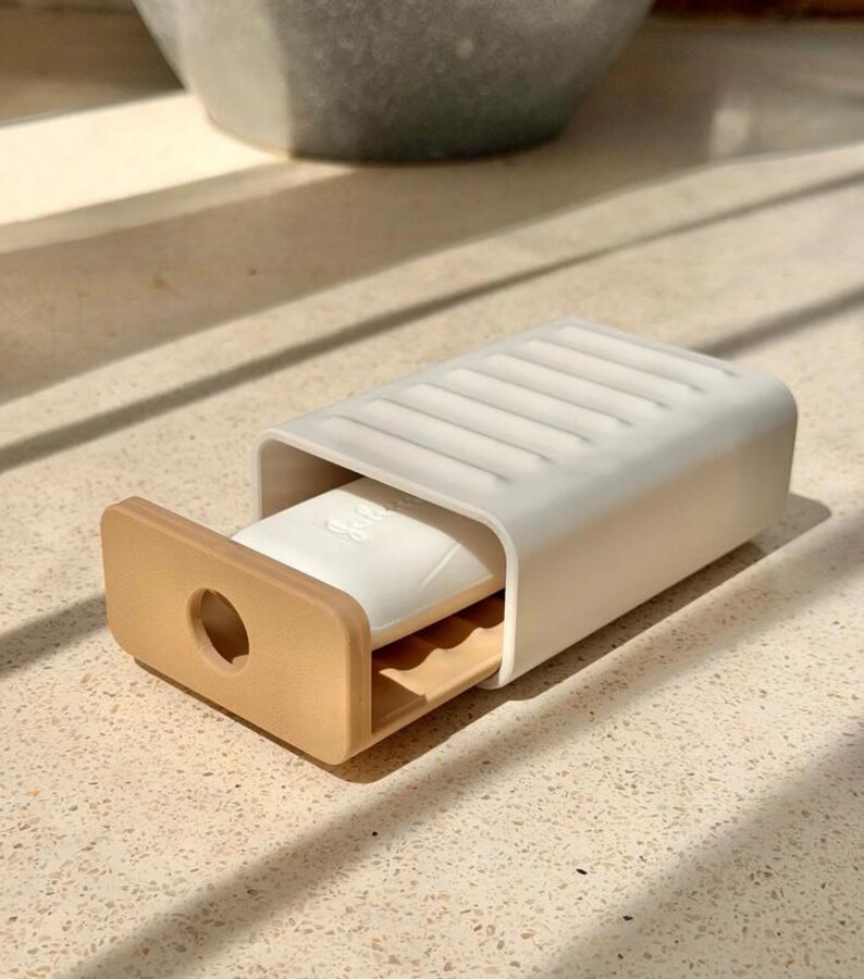May include: A rectangular soap dish featuring a white sliding cover with a ribbed top surface and a tan base tray. The tray includes a circular cutout on the front panel and a white bar of soap with embossed script text resting inside the holder.