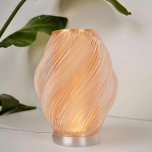 May include: A peach-coloured, swirled glass lamp with a soft glow. The lamp has a rounded base and a textured, spiral design. The lamp is plugged in with a white cord. The background includes green leaves.