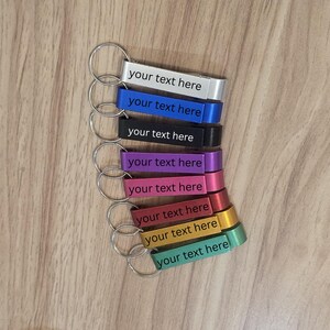 May include: A selection of colourful keychains, including silver, blue, black, purple, pink, red, gold, and green. Each rectangular keychain has the text "your text here". The keychains are attached to silver key rings. The keychains are arranged on a wooden surface.