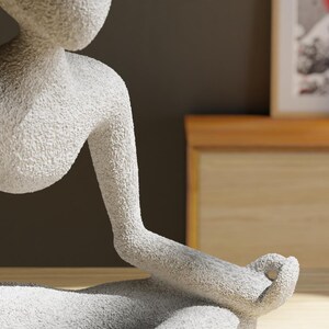 May include: A textured, white sculpture of a person in a seated yoga pose. The figure's arms are extended, with the hands resting in a meditative position. The sculpture is on a wooden surface, with a blurred background.