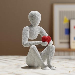 Puede incluir: Escultura gris texturizada de una figura sentada sosteniendo un coraz&oacute;n rojo. La figura tiene un dise&ntilde;o minimalista y est&aacute; colocada sobre una superficie similar al m&aacute;rmol. Arte decorativo para la decoraci&oacute;n del hogar.