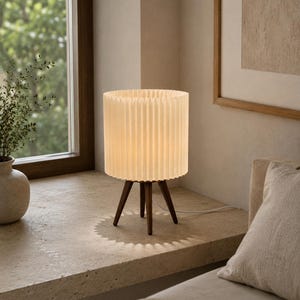 Puede incluir: Una l&aacute;mpara de mesa con una pantalla cil&iacute;ndrica plisada de color blanco roto, sostenida por tres patas de madera de color marr&oacute;n oscuro. La l&aacute;mpara emite una luz c&aacute;lida y suave, proyectando una sombra en forma de sol sobre la superficie.