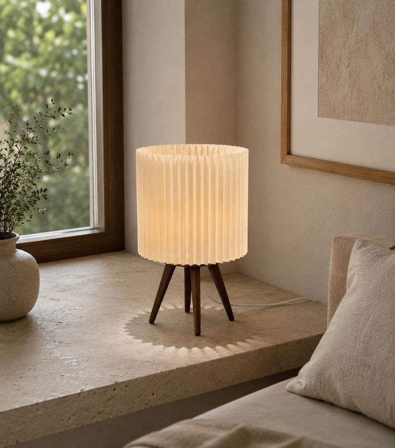 Puede incluir: Una l&aacute;mpara de mesa con una pantalla cil&iacute;ndrica plisada de color blanco roto, sostenida por tres patas de madera de color marr&oacute;n oscuro. La l&aacute;mpara emite una luz c&aacute;lida y suave, proyectando una sombra en forma de sol sobre la superficie.