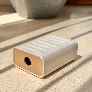 May include: A rectangular white soap dish featuring a ribbed top surface and a solid tan-colored end panel with a circular hole in the center. The object has a smooth, matte finish and a clean, geometric design with rounded edges on all sides.
