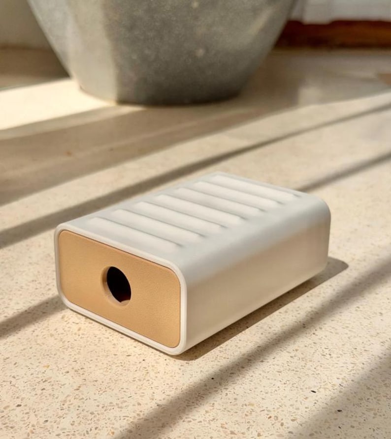 May include: A rectangular white soap dish featuring a ribbed top surface and a solid tan-colored end panel with a circular hole in the center. The object has a smooth, matte finish and a clean, geometric design with rounded edges on all sides.