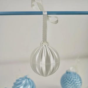 Pode incluir: Um ornamento esf&eacute;rico branco brilhante com sulcos verticais, suspenso por uma fita de cetim branca numa haste horizontal azul. O ornamento apresenta uma superf&iacute;cie lisa e refletora com um pequeno la&ccedil;o no topo para pendurar.
