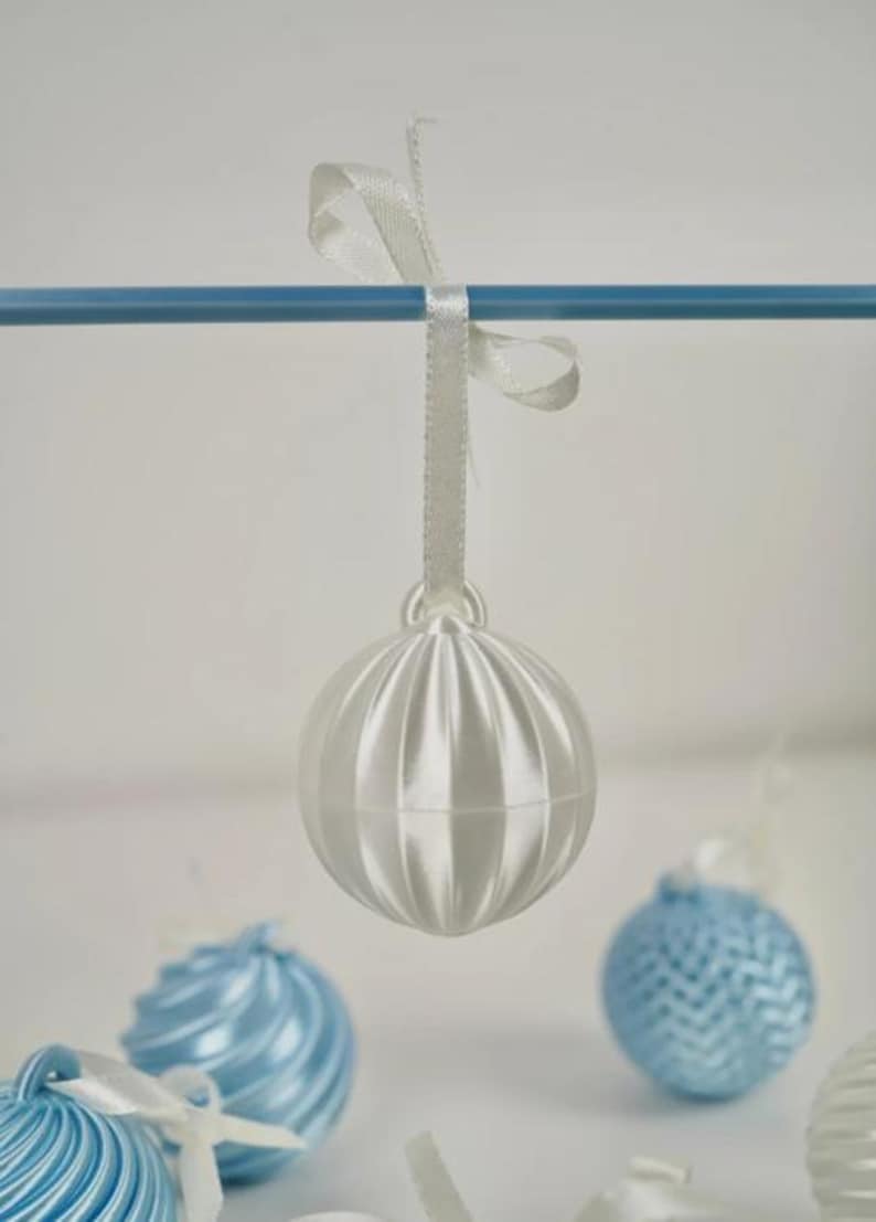 Pode incluir: Um ornamento esf&eacute;rico branco brilhante com sulcos verticais, suspenso por uma fita de cetim branca numa haste horizontal azul. O ornamento apresenta uma superf&iacute;cie lisa e refletora com um pequeno la&ccedil;o no topo para pendurar.