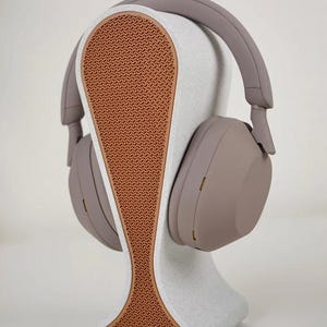 May include: A pair of taupe-colored over-ear headphones resting on a white and brown headphone stand. The stand has a textured brown center and a curved design. The headphones have a sleek, modern aesthetic.