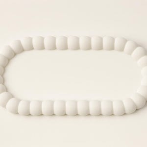May include: A white, oval-shaped decorative object. The object is composed of a series of rounded, connected segments, creating a textured, beaded appearance. The design is simple and minimalist, with a focus on form and texture.