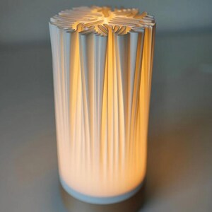 Puede incluir: Una l&aacute;mpara cil&iacute;ndrica blanca con un dise&ntilde;o plisado texturizado. La parte superior tiene un patr&oacute;n calado decorativo. La l&aacute;mpara emite un brillo amarillo c&aacute;lido desde el interior y se asienta sobre una base dorada. Un art&iacute;culo de decoraci&oacute;n del hogar moderno.