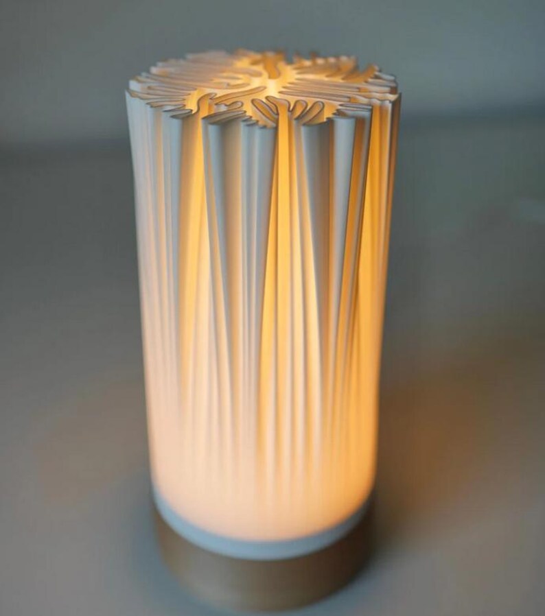 Puede incluir: Una l&aacute;mpara cil&iacute;ndrica blanca con un dise&ntilde;o plisado texturizado. La parte superior tiene un patr&oacute;n calado decorativo. La l&aacute;mpara emite un brillo amarillo c&aacute;lido desde el interior y se asienta sobre una base dorada. Un art&iacute;culo de decoraci&oacute;n del hogar moderno.