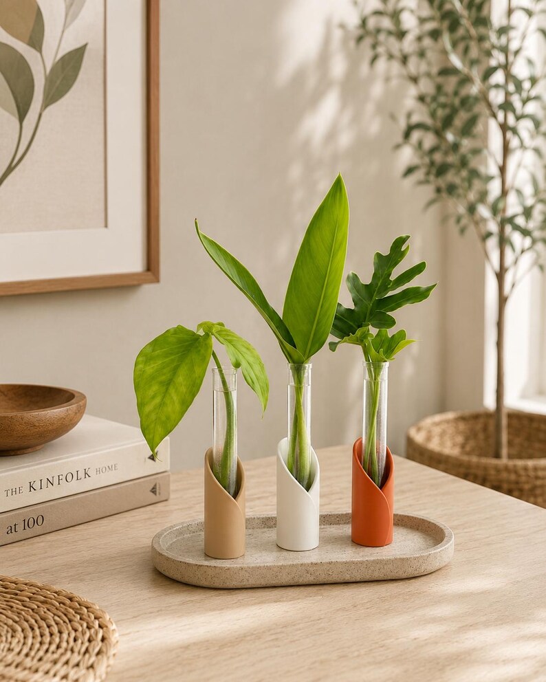 May include: Three clear glass test tube vases held in cylindrical stands of beige, white, and terracotta colours, arranged on an oval speckled tray. Each vase contains a single green plant cutting with distinct leaf shapes and sizes.