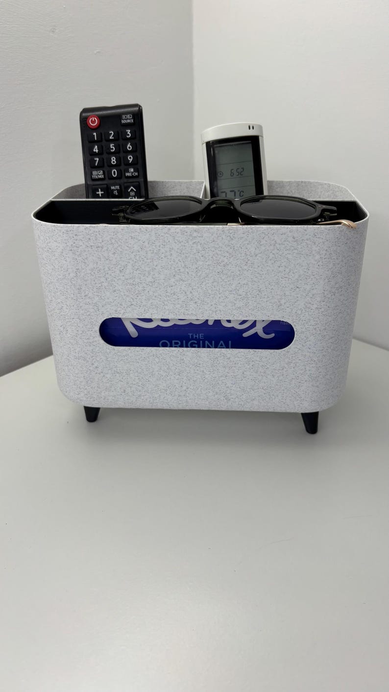 Op de afbeelding: Een grijze, gespikkelde tissuebox met een rechthoekige opening, met een blauw-witte tissuebox. Een zwarte afstandsbediening, een klein wit apparaat en een zwarte zonnebril liggen bovenop. De tissuebox heeft vier zwarte poten.