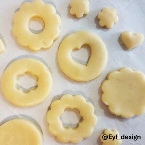 May include: Unbaked light beige cookie dough cut into various shapes including scalloped circles with center cutouts, a circle with a heart cutout, a solid heart, and small flower shapes. The text Eyf_design is visible in the bottom right corner.